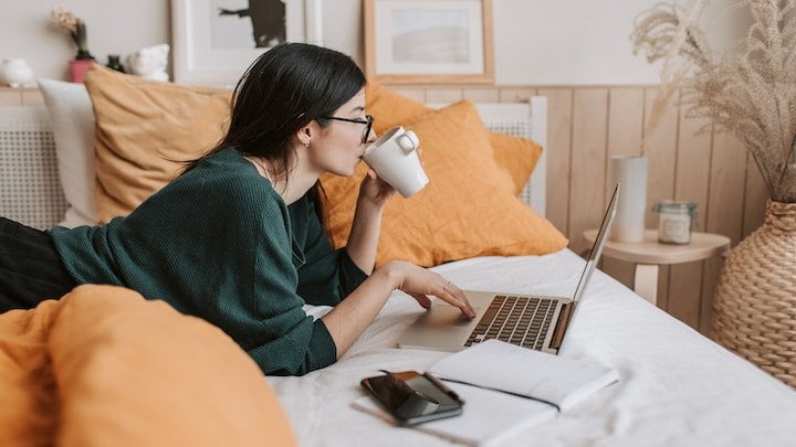 Woman drinking a hot drink and typing on a laptop in bed at a residential drug and alcohol rehab in Norfolk